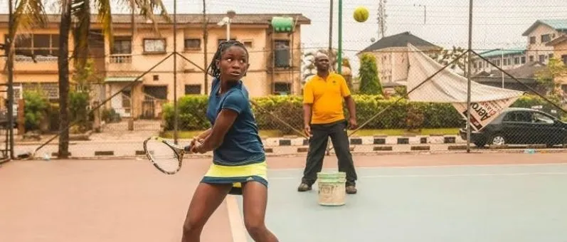 A young Nigerian girl hits a backhand on a Lagos court while her coach feeds balls from a bucket